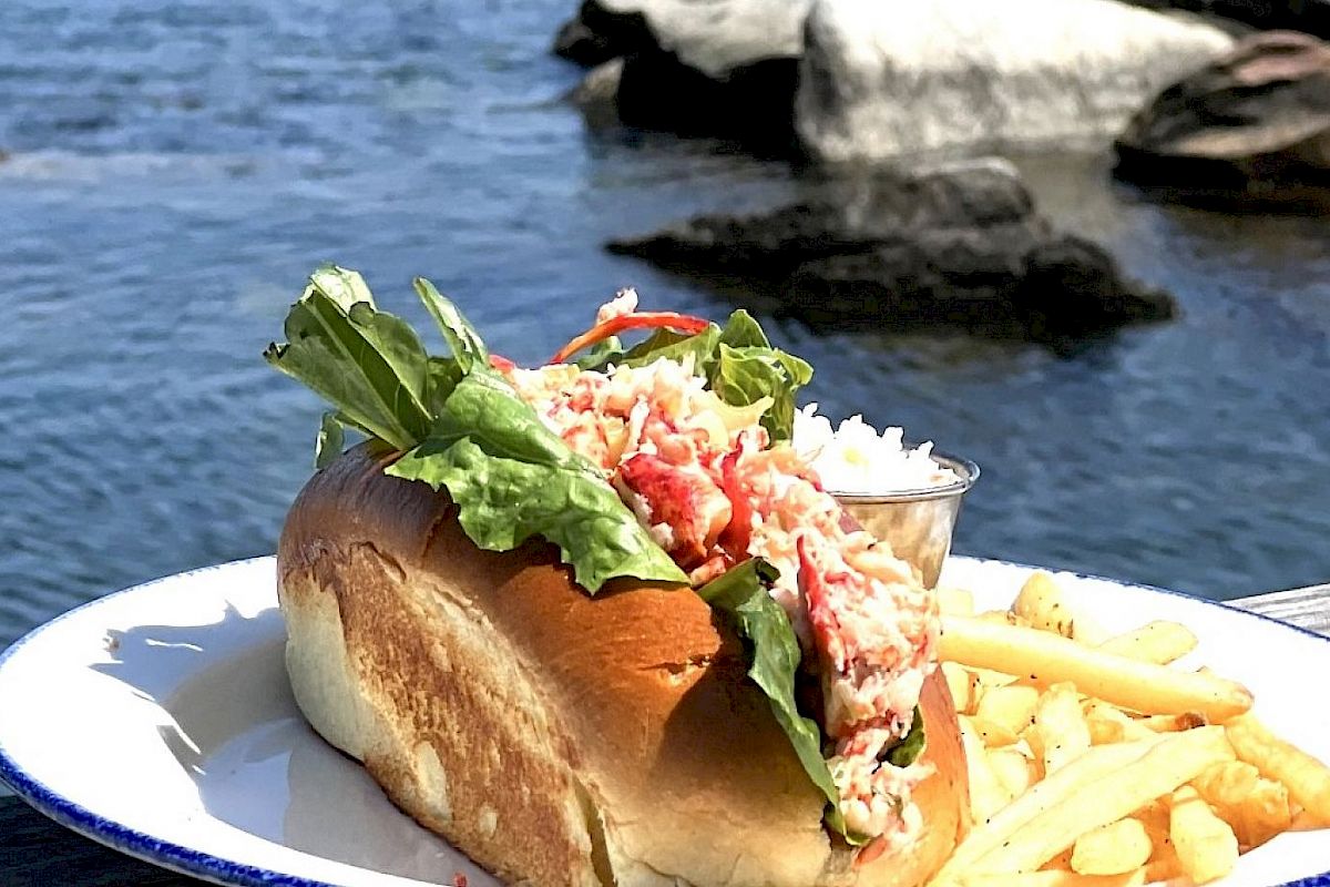 A lobster roll with lettuce, a side of fries, and coleslaw on a plate, set on a wooden table next to a body of water.