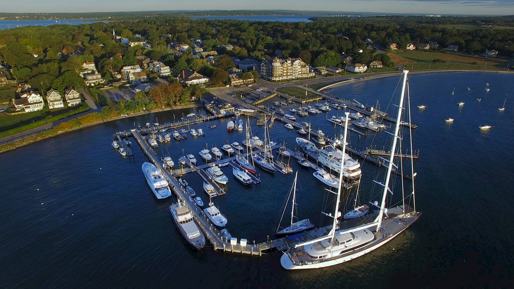 An aerial view of a marina with multiple yachts and boats docked, surrounded by a coastal town with houses and greenery in the background.