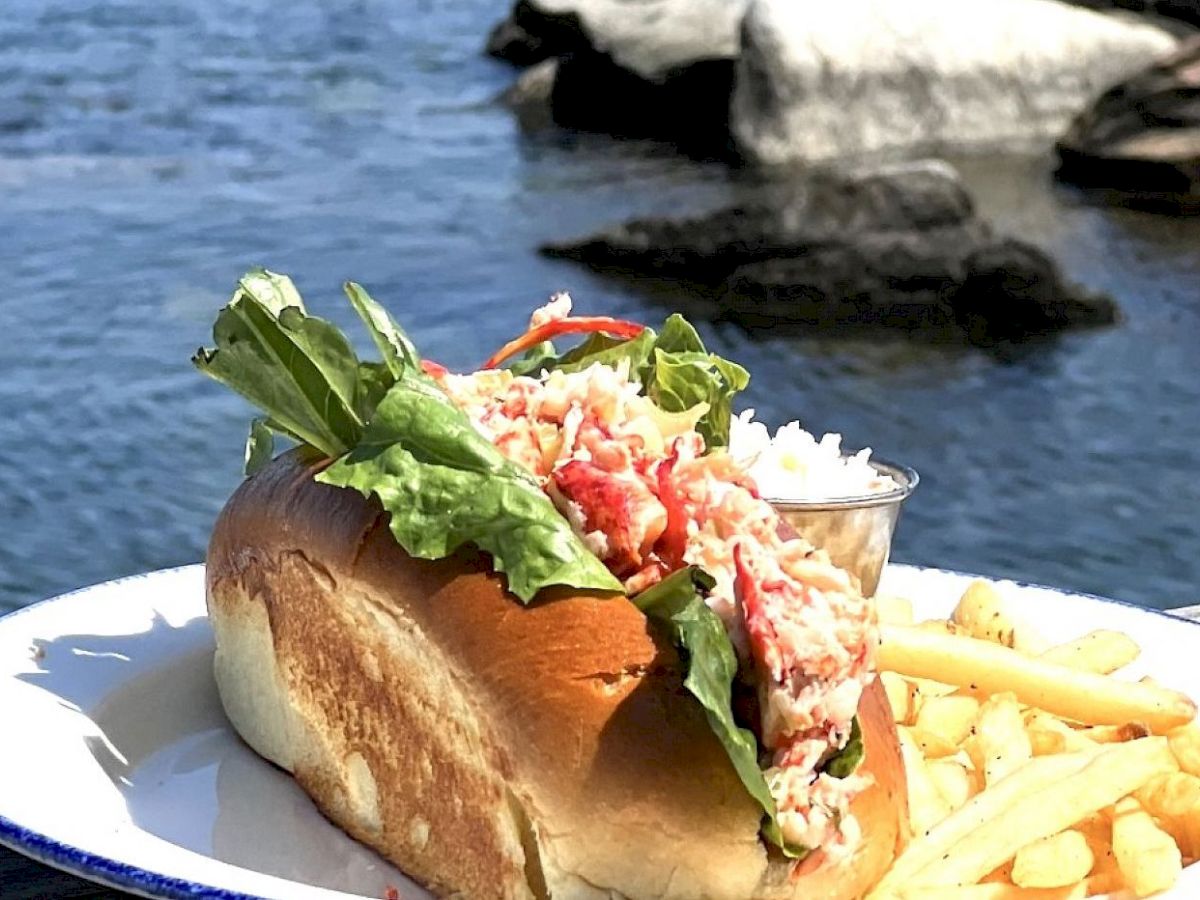 A lobster roll with lettuce, a side of fries, and coleslaw on a plate, set on a wooden table next to a body of water.