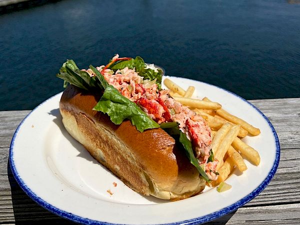 The image shows a lobster roll with lettuce on a plate, accompanied by French fries, by the waterfront.