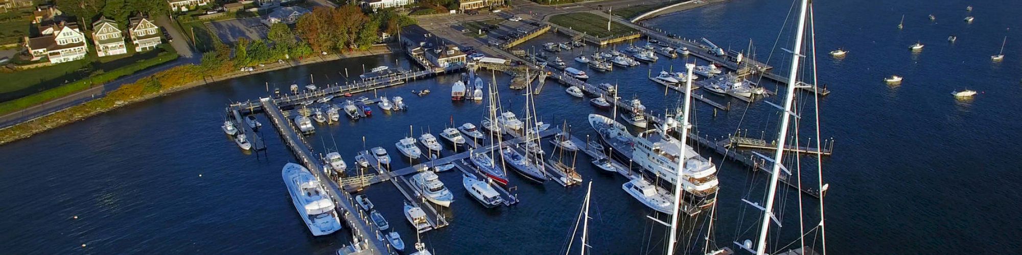 An aerial view of a marina with several yachts and boats docked, surrounded by a coastal town and lush greenery, with a distant water body visible.