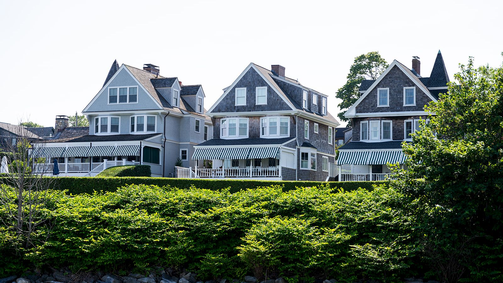 Cozy row of seaside houses with blue-gray siding, peaked roofs, and wraparound porches perched above a green hedge along the shore.