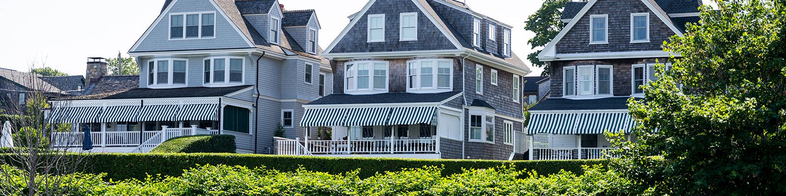 Cozy row of seaside houses with blue-gray siding, peaked roofs, and wraparound porches perched above a green hedge along the shore.