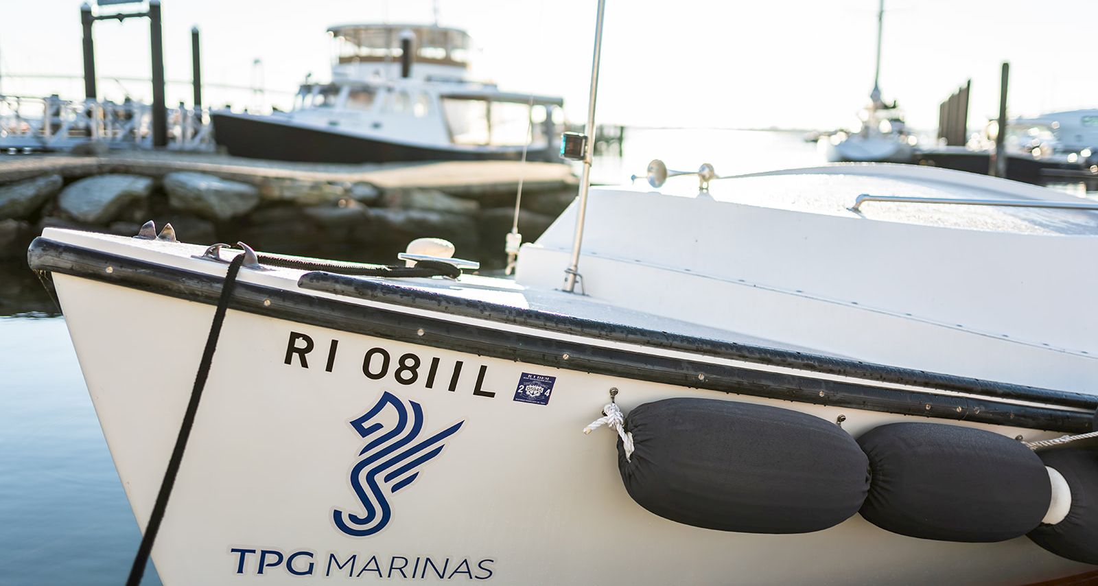 A white boat docked at a marina with &ldquo;TPG MARINAS&rdquo; logo and registration RI 0811L on the hull, calm water and other boats in the background.