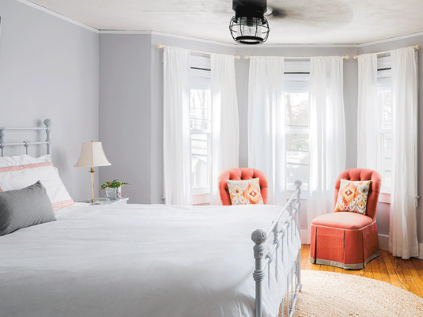 A bright, airy bedroom with a white metal bed, soft gray walls, sheer white curtains, and two coral armchairs by the window.