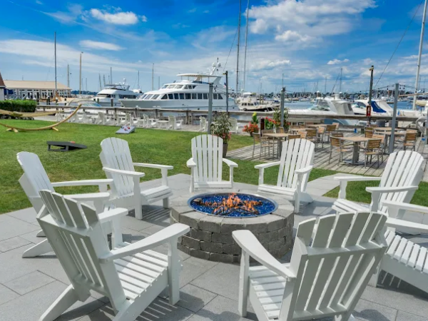 A marina backdrop with several white Adirondack chairs around a stone fire pit, blue sky, and yachts anchored in the harbor.