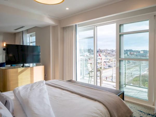 Hotel bedroom with a large bed, TV on a wooden dresser, glass doors to a balcony, and city views outside.
