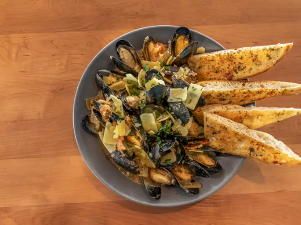 A plate of mussels with roasted vegetables and long pita wedges, served on a round gray dish on a wooden table.
