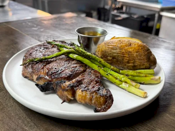 Juicy grilled steak with a charred crust, tender center, roasted potato, and grilled asparagus, served with a au jus cup on the plate.