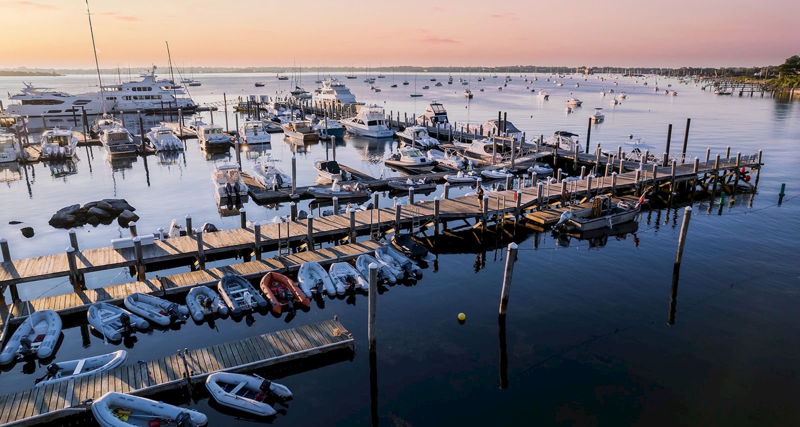 Boats and a long wooden pier at a calm marina at sunset, with calm water reflecting pink skies and several small boats docked along the pier.