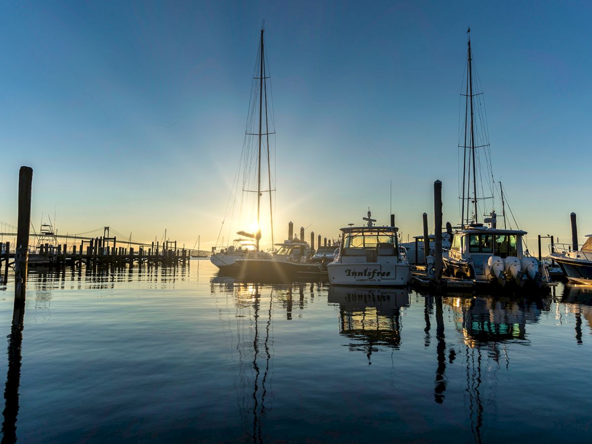 Boats docked at a calm harbor with masts reflecting on the glassy water at sunset, a peaceful marina scene with wooden piers.