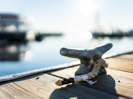 A rusty mooring cleat on a wooden dock with a blurred marina and glimmering water in the background, casting a long shadow.