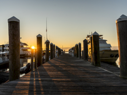 A wooden pier extends into the water at sunset, with boats on either side and long shadows, calm sky turning warm orange near the horizon.