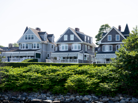 A row of large coastal houses with gabled roofs sits on a grassy hill above a stone-lined shoreline.