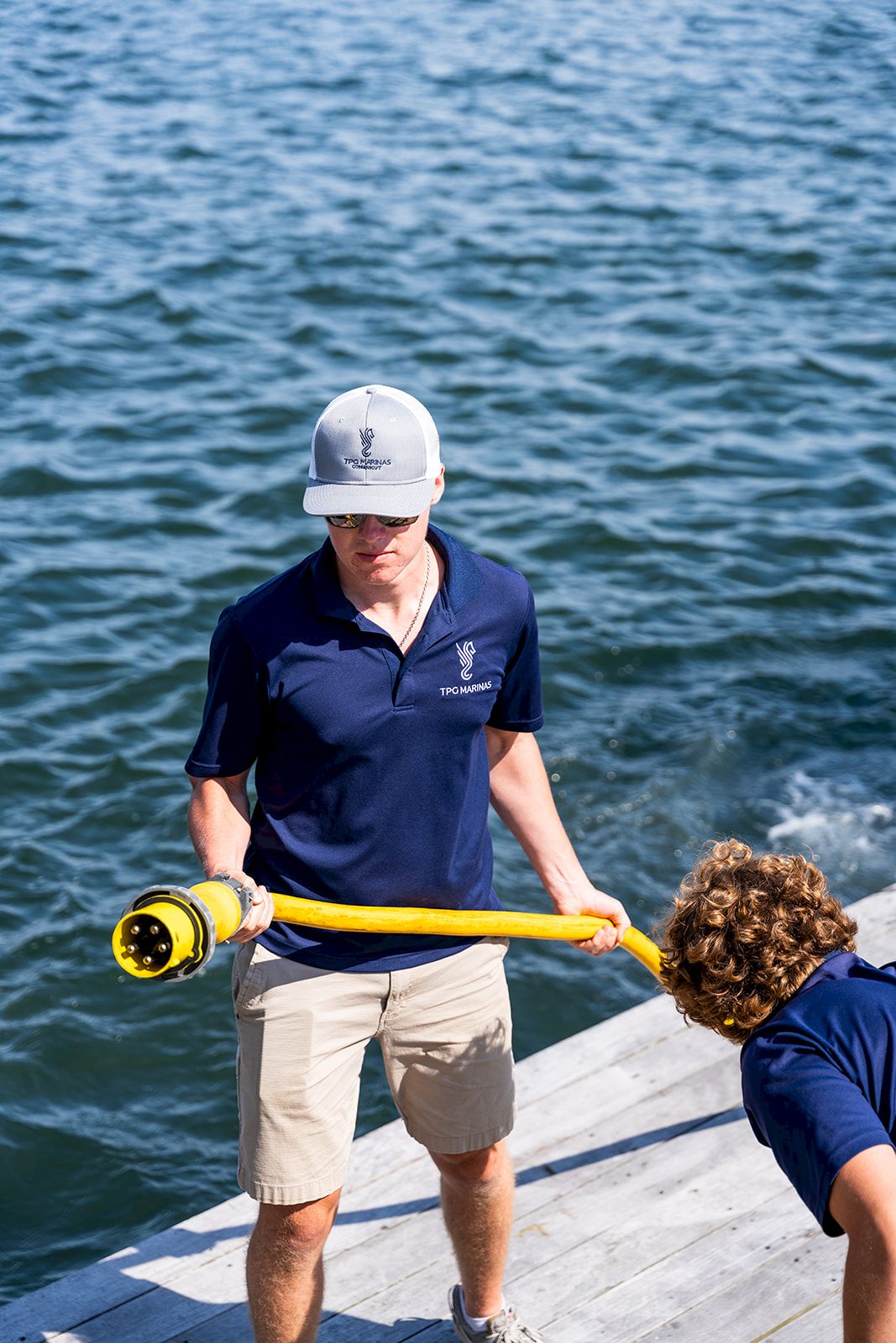 A man in a navy polo and beige shorts holds a yellow water-sampling tool on a dock by blue-green water, with another person bent over nearby.