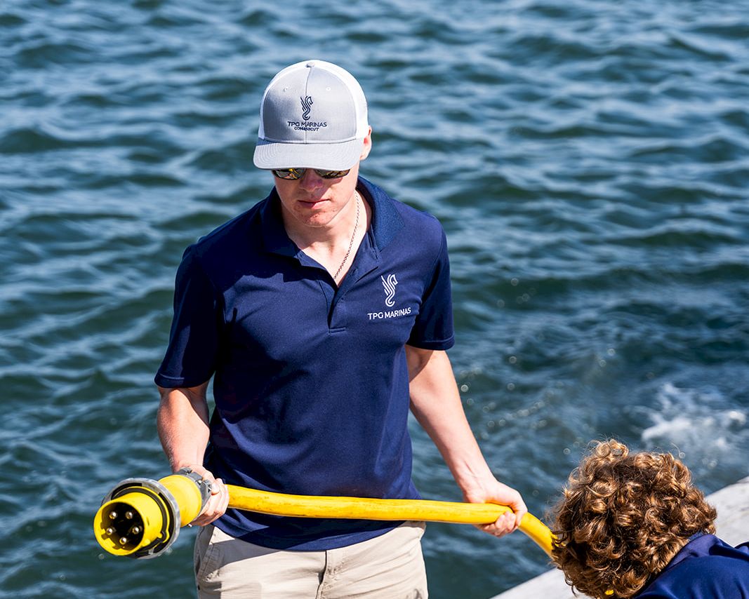 A man in a navy polo and beige shorts holds a yellow water-sampling tool on a dock by blue-green water, with another person bent over nearby.