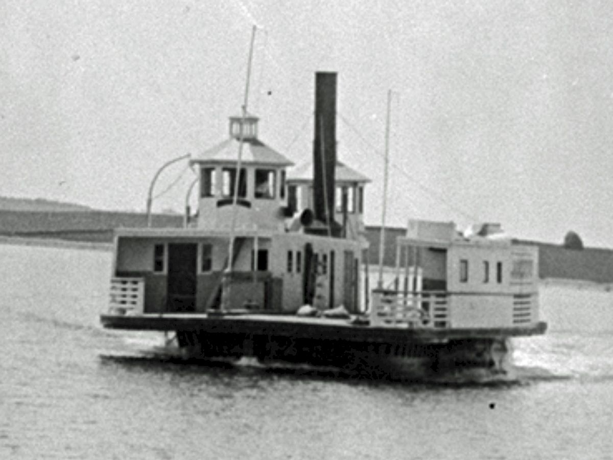 A small vintage steamboat with a tall smokestack, two decks, and a cabin; sits on calm water with flat shoreline in the distance.