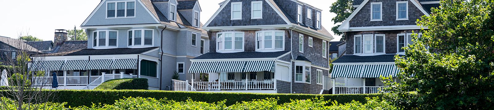 Row of charming grey-blue seaside houses with white trim, a wooden balcony, and manicured hedges along a rocky shore.