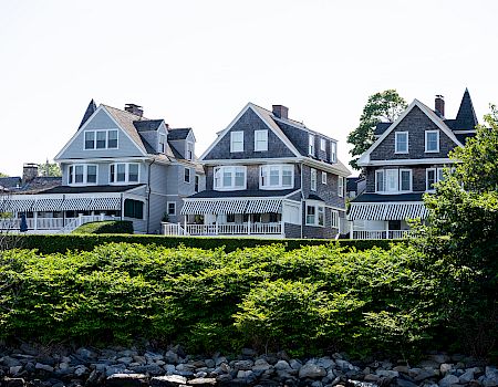 Row of charming grey-blue seaside houses with white trim, a wooden balcony, and manicured hedges along a rocky shore.