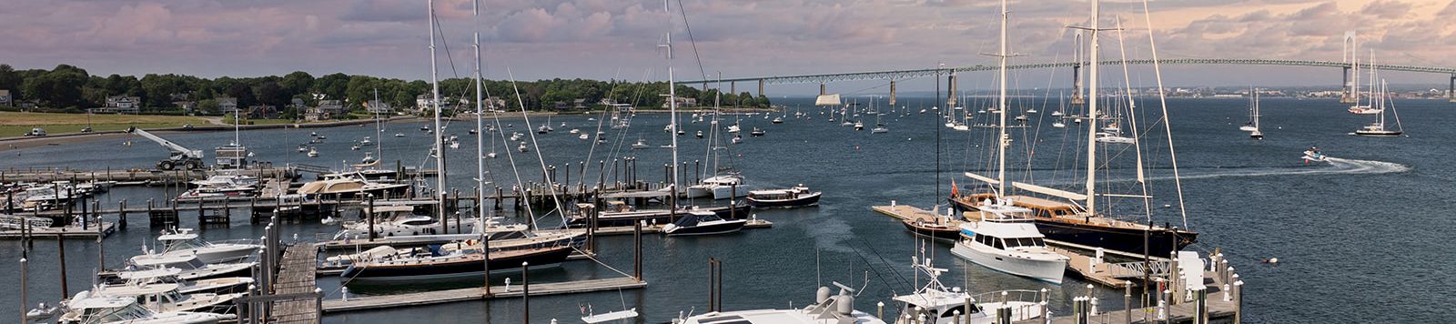 A bustling marina with several docked yachts and sailboats, calm water, and a colorful sunset sky casting pinkish clouds over the scene.