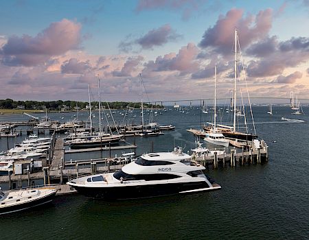 A bustling marina with several docked yachts and sailboats, calm water, and a colorful sunset sky casting pinkish clouds over the scene.