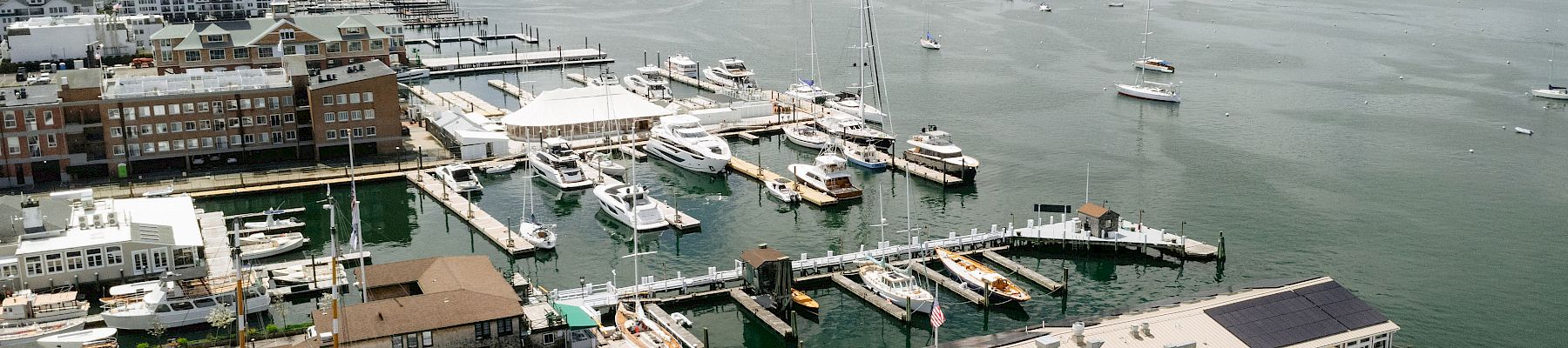 A harbor scene with numerous boats and yachts docked at piers, buildings along the waterfront, calm water, and a distant shoreline.