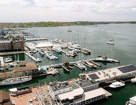 A harbor scene with numerous boats and yachts docked at piers, buildings along the waterfront, calm water, and a distant shoreline.