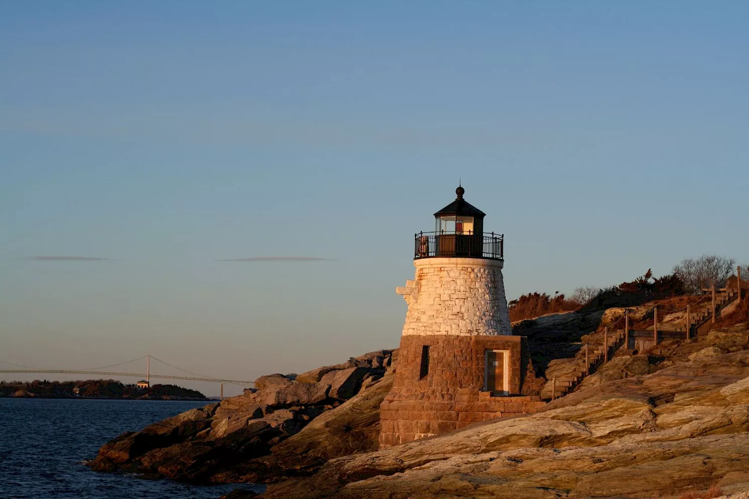 A small lighthouse perched on rocky coastline at sunset, overlooking calm water with clear sky and a stone wall path along the cliffs.