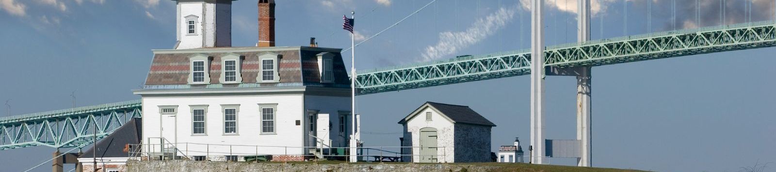 A lighthouse on a rocky coast with a small white building, connected by a bridge to mainland, under a partly cloudy sky.