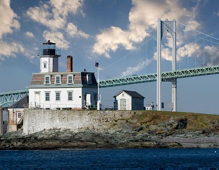A lighthouse on a rocky coast with a small white building, connected by a bridge to mainland, under a partly cloudy sky.