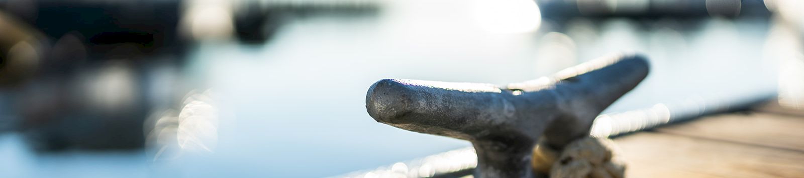 A weathered boat cleat on a wooden dock, with a blurred marina in the bright background, sunlight reflecting on calm water.