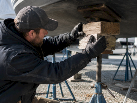 A man in a cap and jacket effects boat hull repairs, wedging wooden blocks under a supported boat on stands.