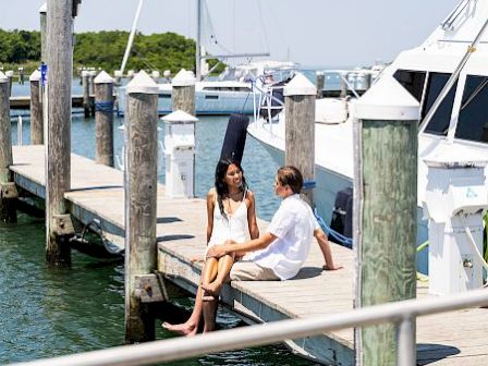 Two people sit on a wooden dock by boats at a marina, chatting and enjoying a sunny day.