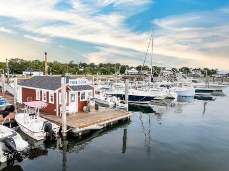 A marina with several boats docked, a small red fuel dock building, calm water, and a bright blue sky with wispy clouds.
