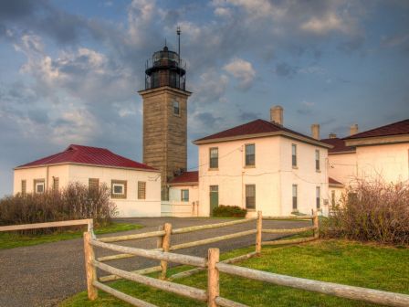 A lighthouse overlooks white and cream keeper houses with red roofs, a wooden fence, and a gravel path under a cloudy sky, serene coastal scene.
