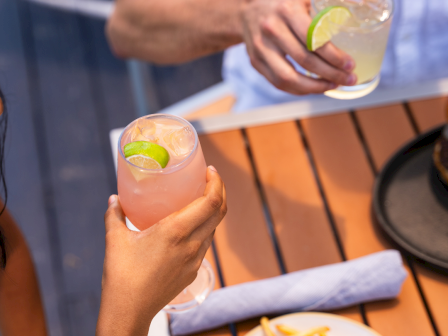 Two people clink drinks with lime wedges on a sunny outdoor table, plus a plate of french fries in the foreground.