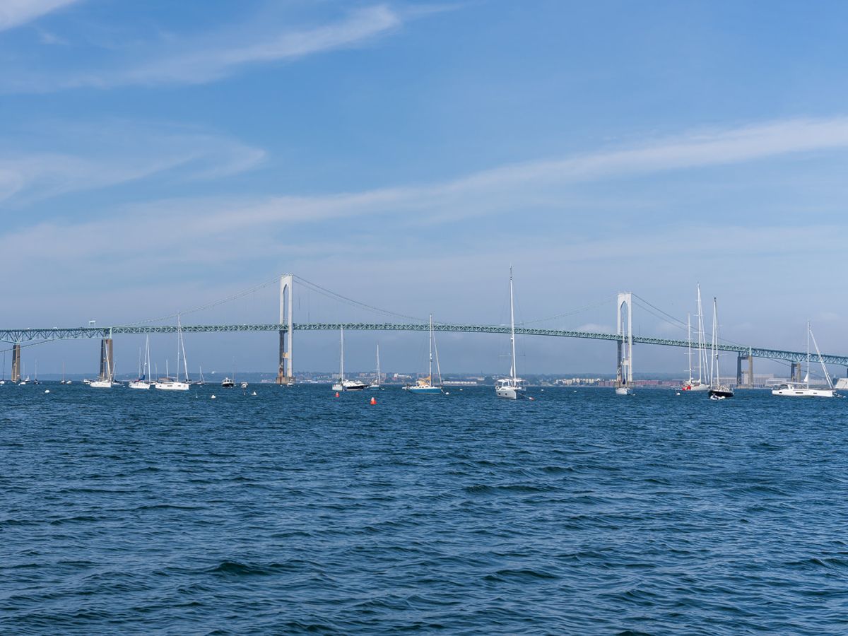 A waterside cityscape with a long bridge and sailboats anchored along the horizon, under a clear blue sky, calm sea, and gentle waves.