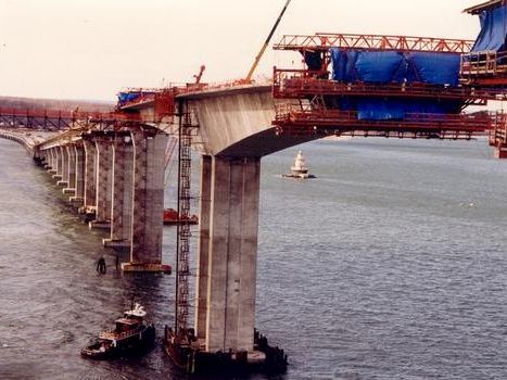A partially built bridge over water with tall concrete piers, construction equipment, and a boat nearby, under clear skies.