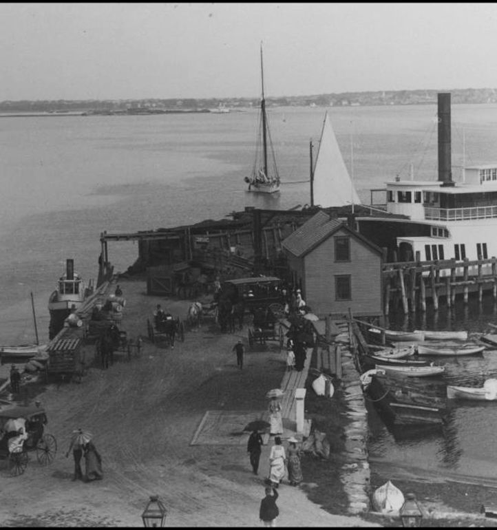 A black-and-white seaside scene: a pier with a large boat, smaller boats, sailboats, a house/boathouse, and people strolling by the water.