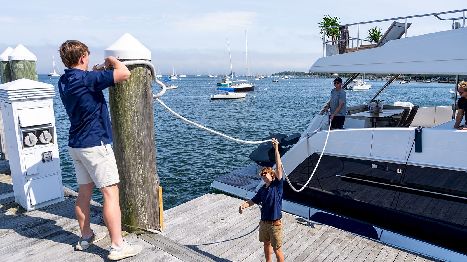 Two people on a marina dock. A man in a blue shirt unties a rope from a post, while a child in a dress holds the other end near a large yacht.