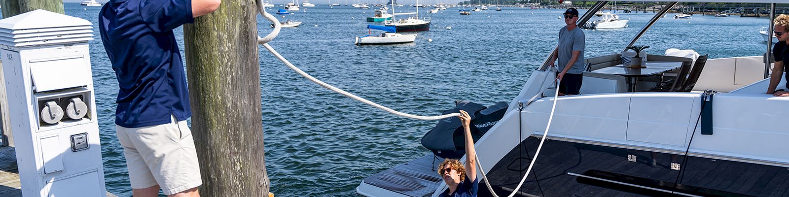 Two people on a marina dock. A man in a blue shirt unties a rope from a post, while a child in a dress holds the other end near a large yacht.