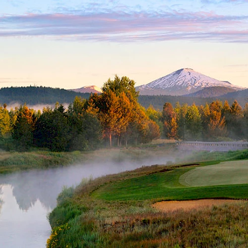 A peaceful golf course scene with a river, lush greenery, trees, and distant snow-capped mountains under a blue sky with light clouds ending the sentence.