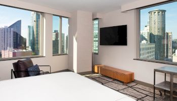 A modern hotel room with city views, a bed, armchair, TV, and table, featuring large windows and contemporary decor.