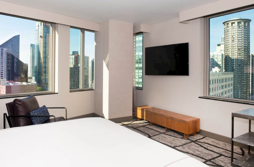 A modern hotel room with city views, a bed, armchair, TV, and table, featuring large windows and contemporary decor.