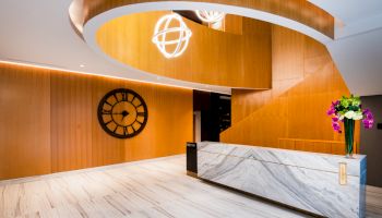 A modern interior with a marble reception desk, wooden walls, a large clock, and unique lighting fixtures; flowers decorate the counter.