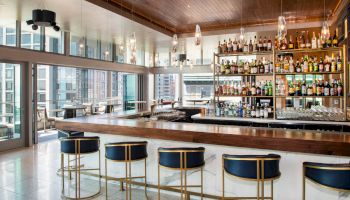 A modern bar with a wooden ceiling, stocked shelves of liquor bottles, stylish blue bar stools, and large windows offering a city view.
