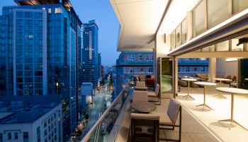 A modern cityscape at dusk with a view of tall buildings from a sleek balcony; includes outdoor seating and a well-lit interior open to the balcony.