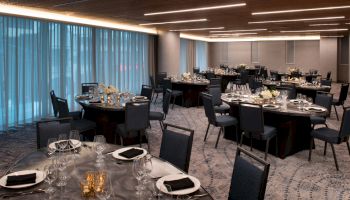 The image shows a modern banquet hall with round tables set for an event, featuring elegant table settings, black napkins, and dim lighting.