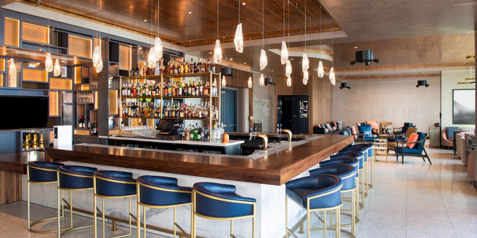 A modern bar with a wooden counter, blue bar stools, hanging lights, and a wide selection of bottles on shelves behind the counter.