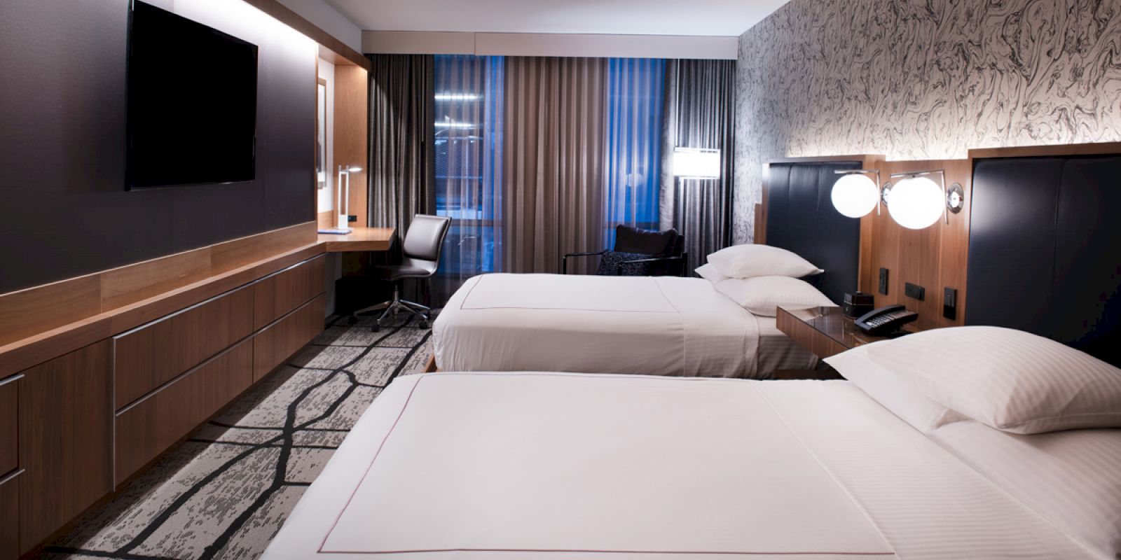 A modern hotel room with two neatly made beds, a wall-mounted TV, a desk with a chair, and window drapes partially drawn, is shown.